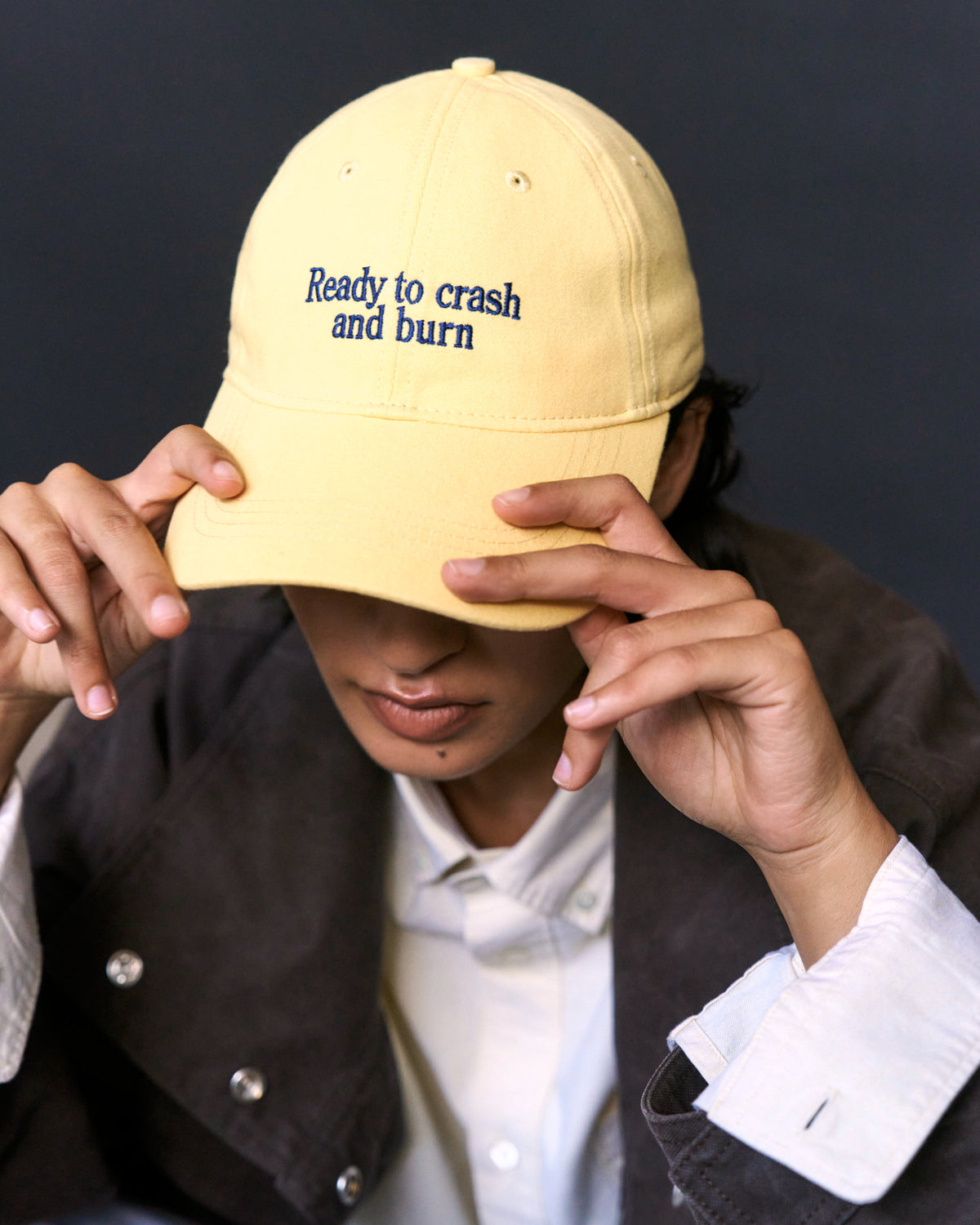 Man pulling the butter yellow Artless Ready To Crash And Burn organic cotton dad cap down over his eyes, brown jacket and white shirt