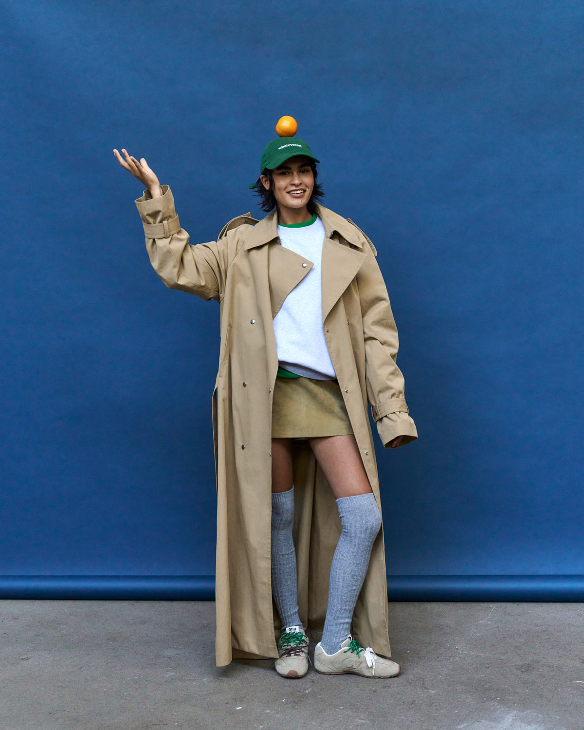 Woman with an arm raised against a blue backdrop wearing the green Artless Whateverest organic cotton dad cap and a beige trench coat