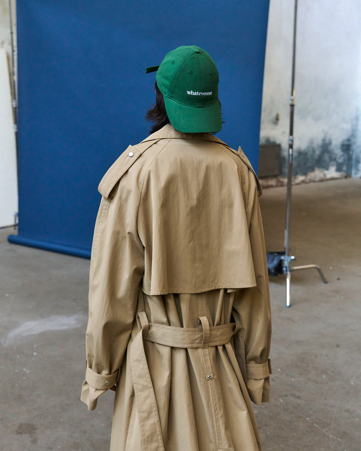 Person in a tan trench coat wearing the green Artless Whateverest organic cotton dad cap seen from the side against a blue backdrop