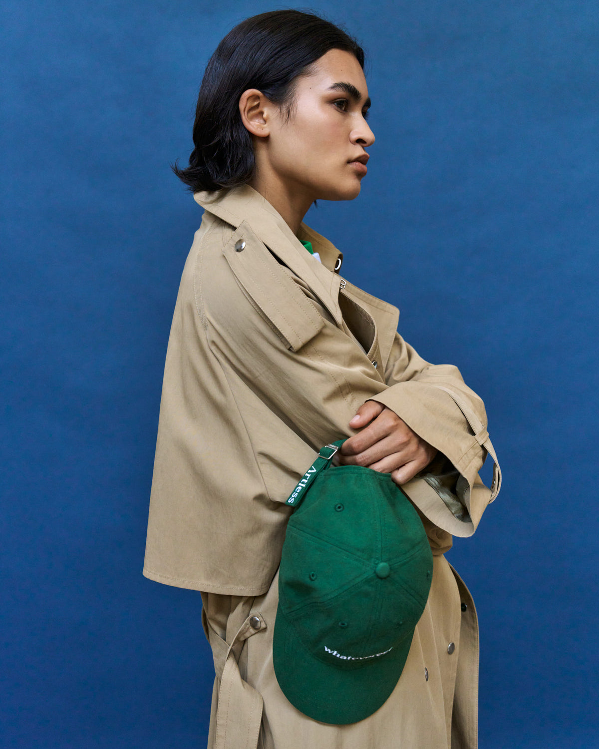 Person in a tan trench coat wearing the green Artless Whateverest organic cotton dad cap against a blue backdrop