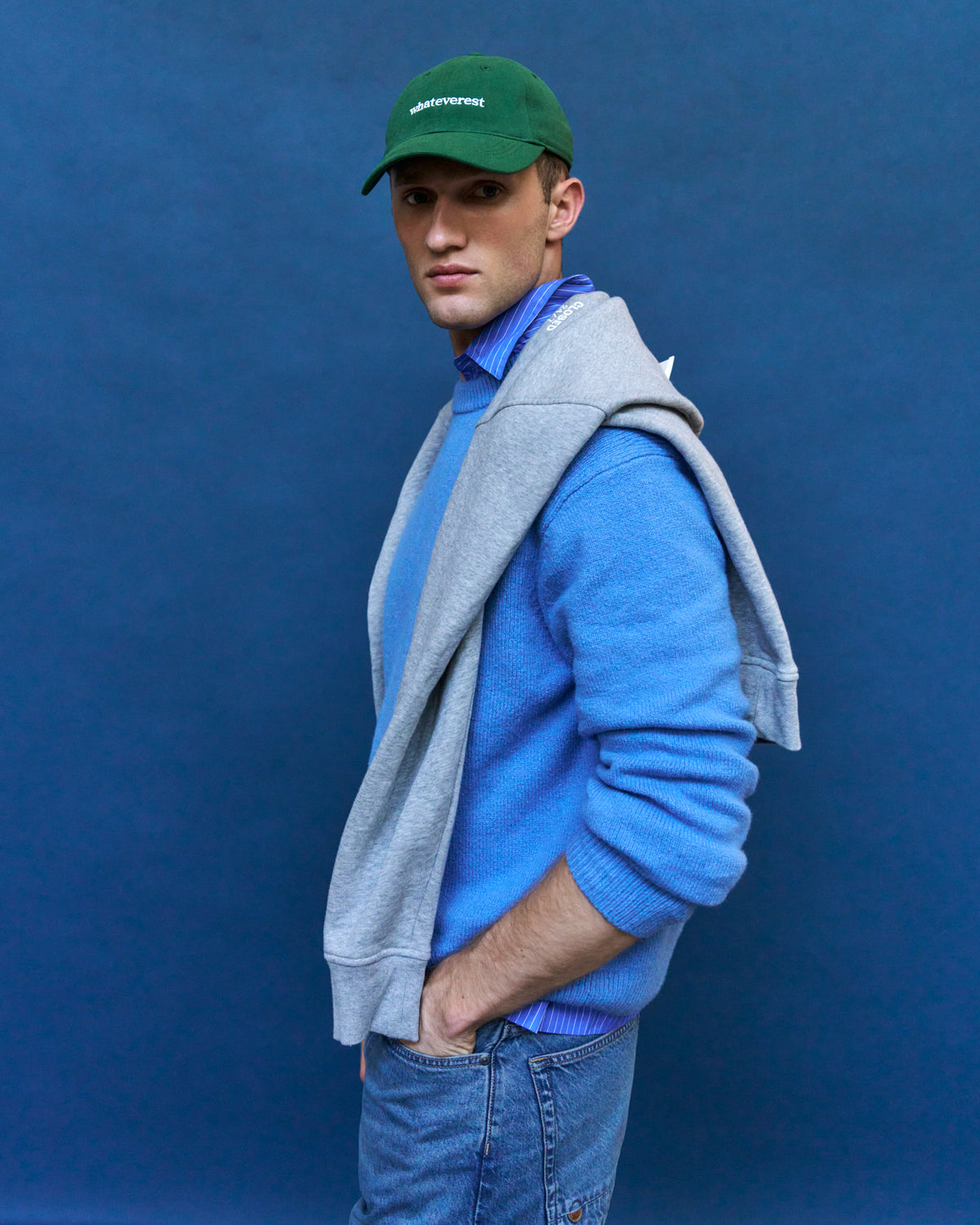 Man in profile against a blue backdrop wearing the green Artless Whateverest organic cotton dad cap, grey sweater and blue fleece