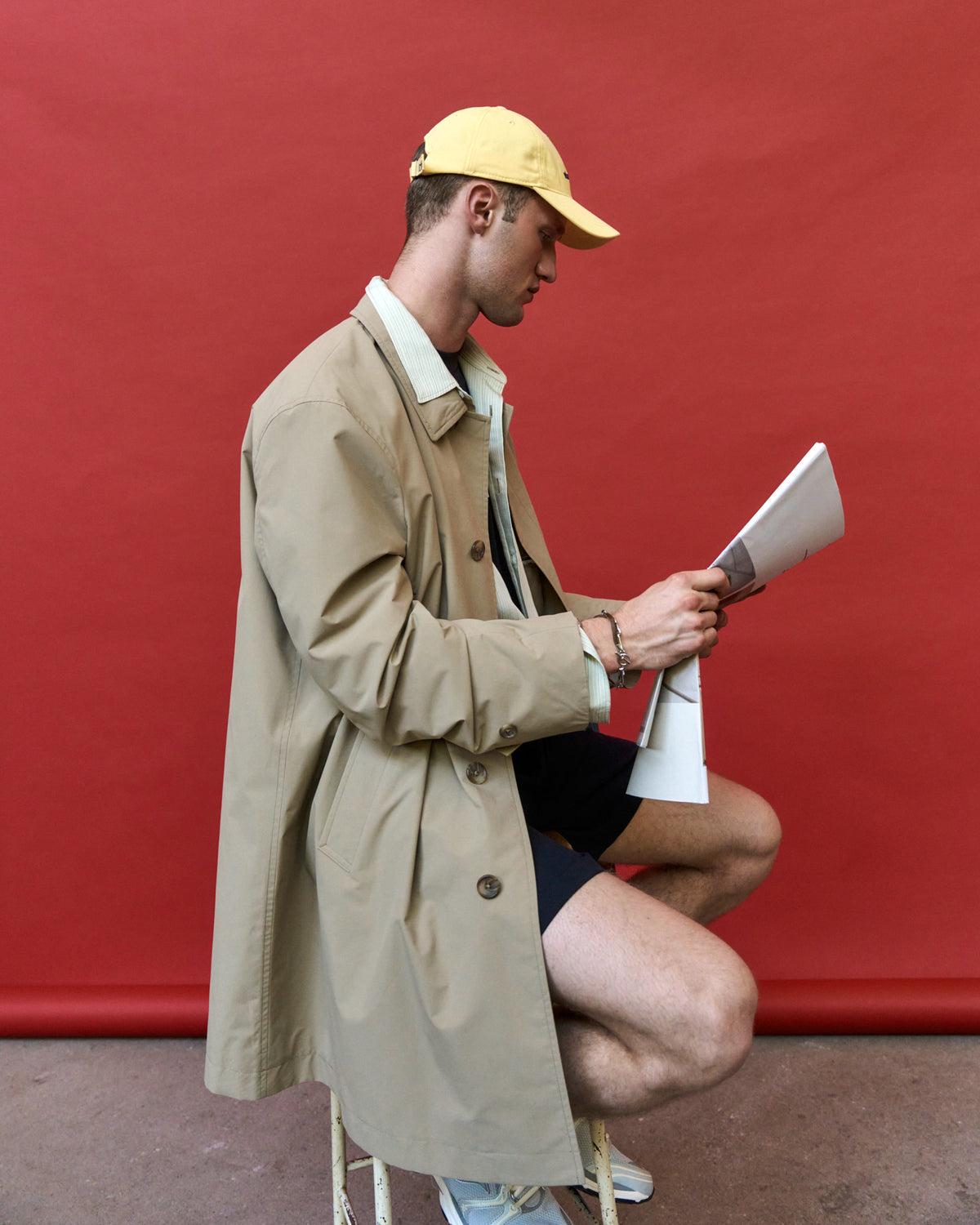 Person crouching against a red wall wearing the butter yellow Artless Whateverest organic cotton dad cap and a beige trench coat