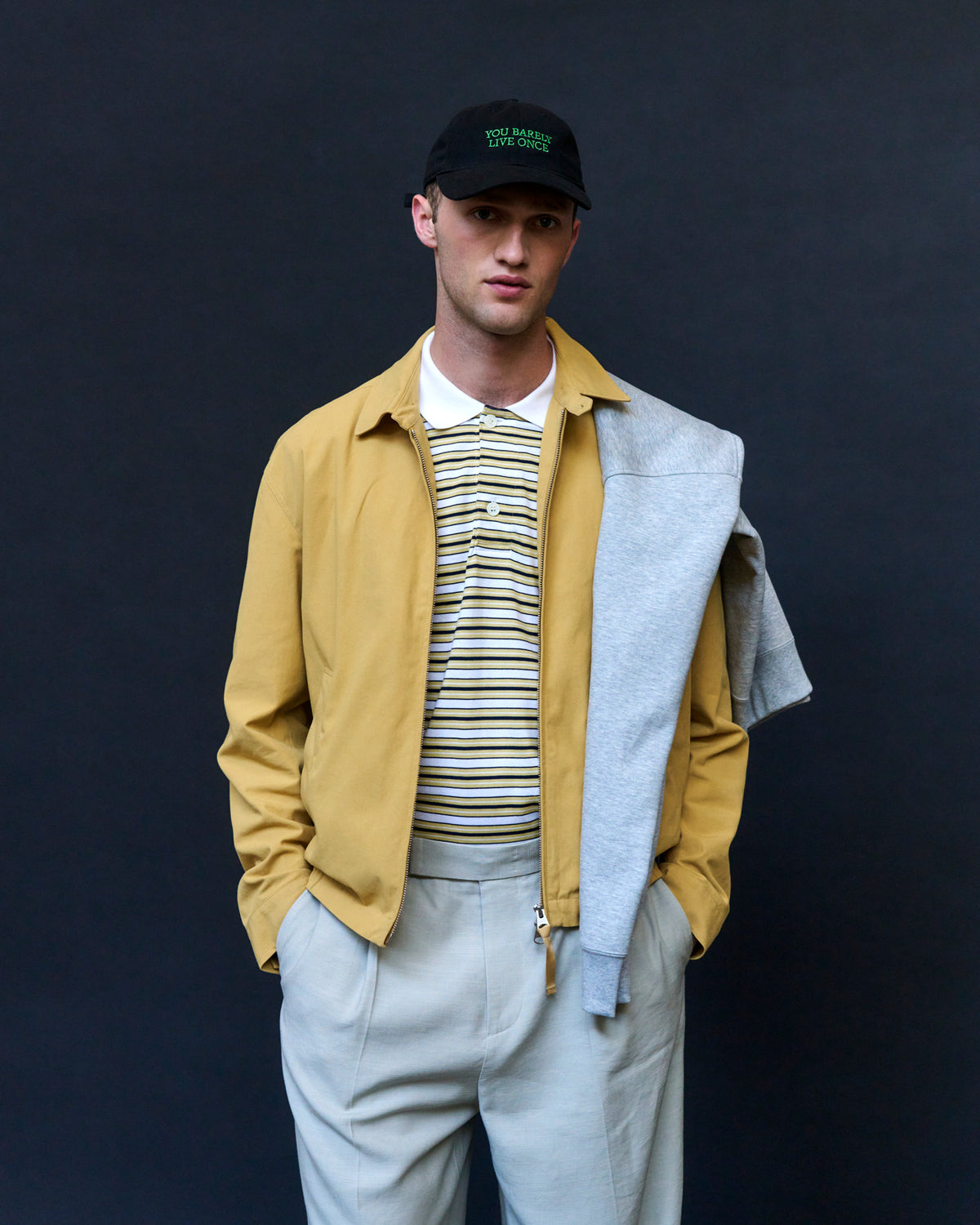 Man against a dark backdrop wearing the black Artless You Barely Live Once organic cotton dad cap, yellow jacket over a striped polo