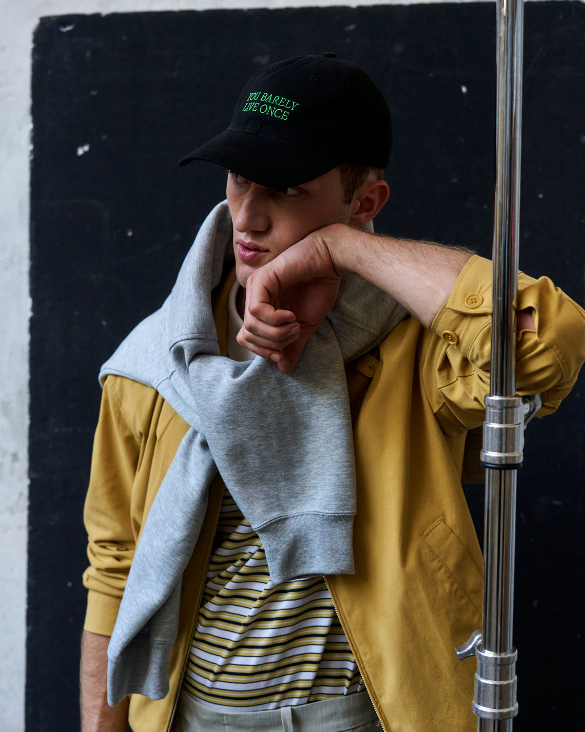 Person seated wearing the black Artless You Barely Live Once organic cotton dad cap, grey hoodie under a yellow jacket
