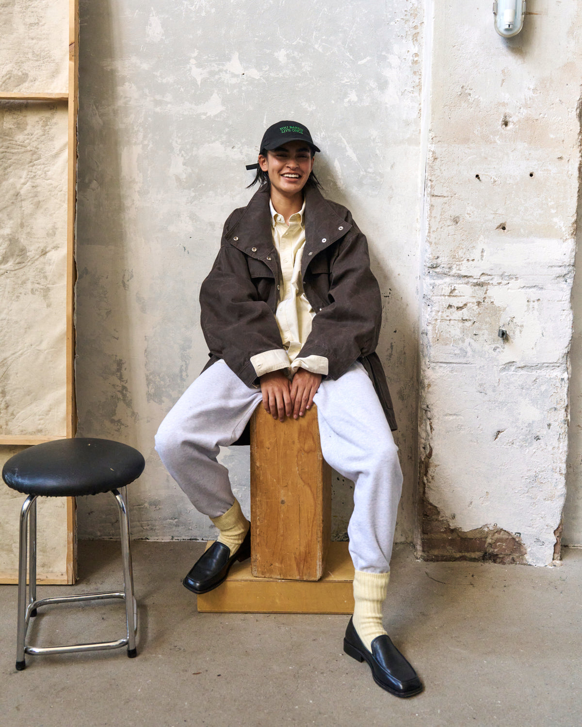 Person seated on a box against a plaster wall wearing the black Artless You Barely Live Once organic cotton dad cap and a brown jacket