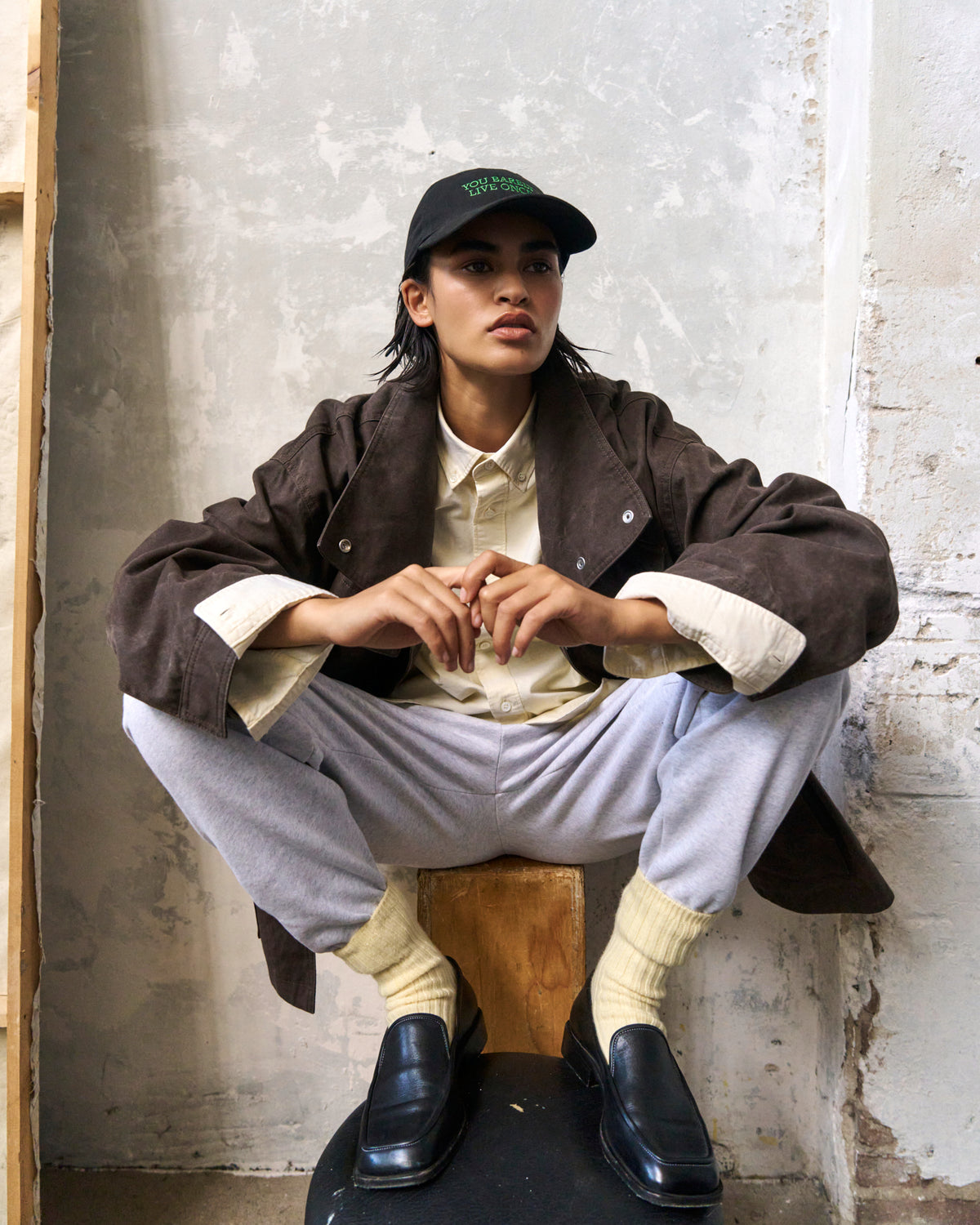 Person seated on a block against a plaster wall wearing the black Artless You Barely Live Once organic cotton dad cap and a brown jacket