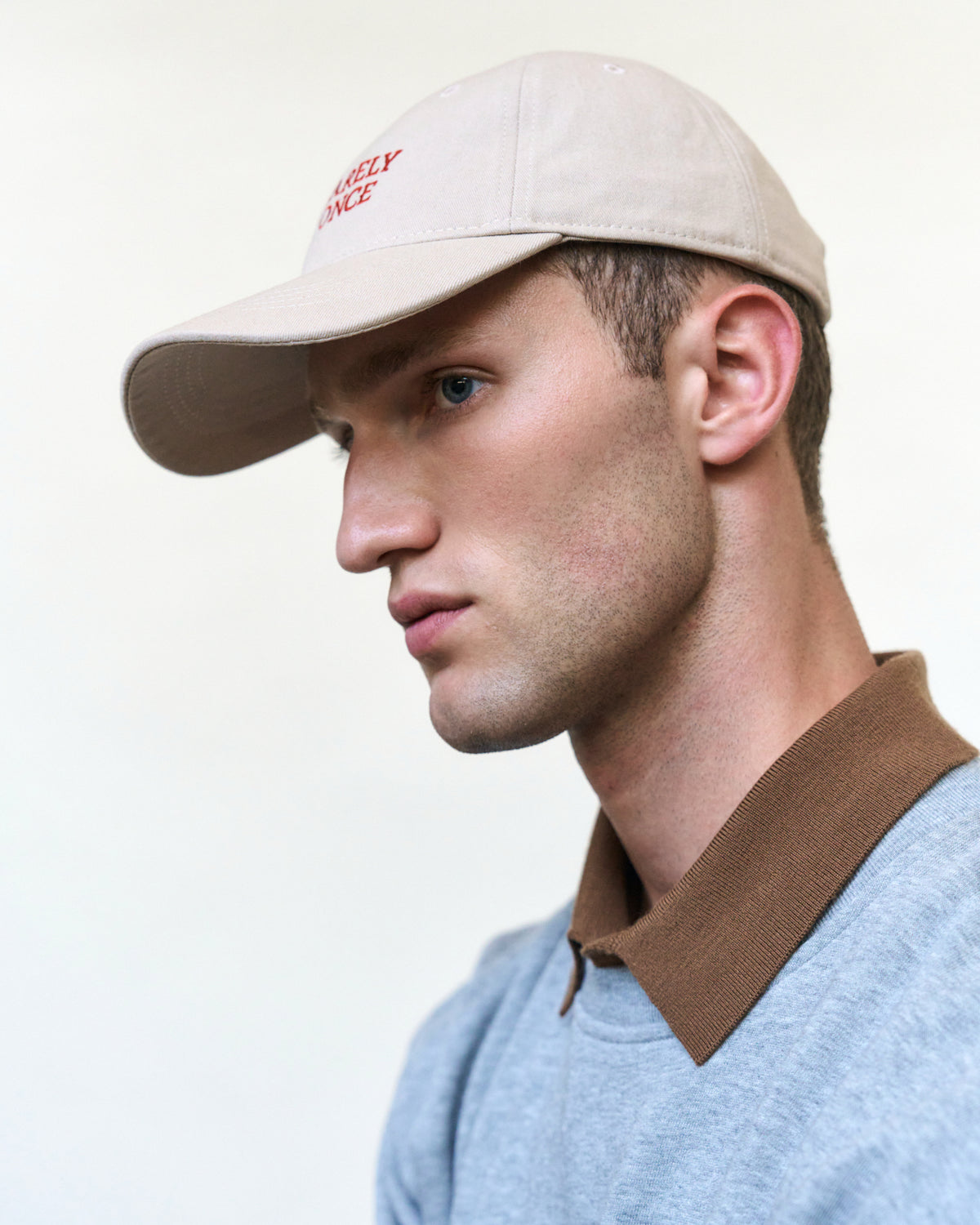 Portrait of a man in profile wearing the sand Artless You Barely Live Once organic cotton dad cap with red embroidery and a grey polo